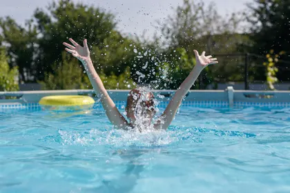 A child playing in a clear backyard pool, arms raised, mid-splash